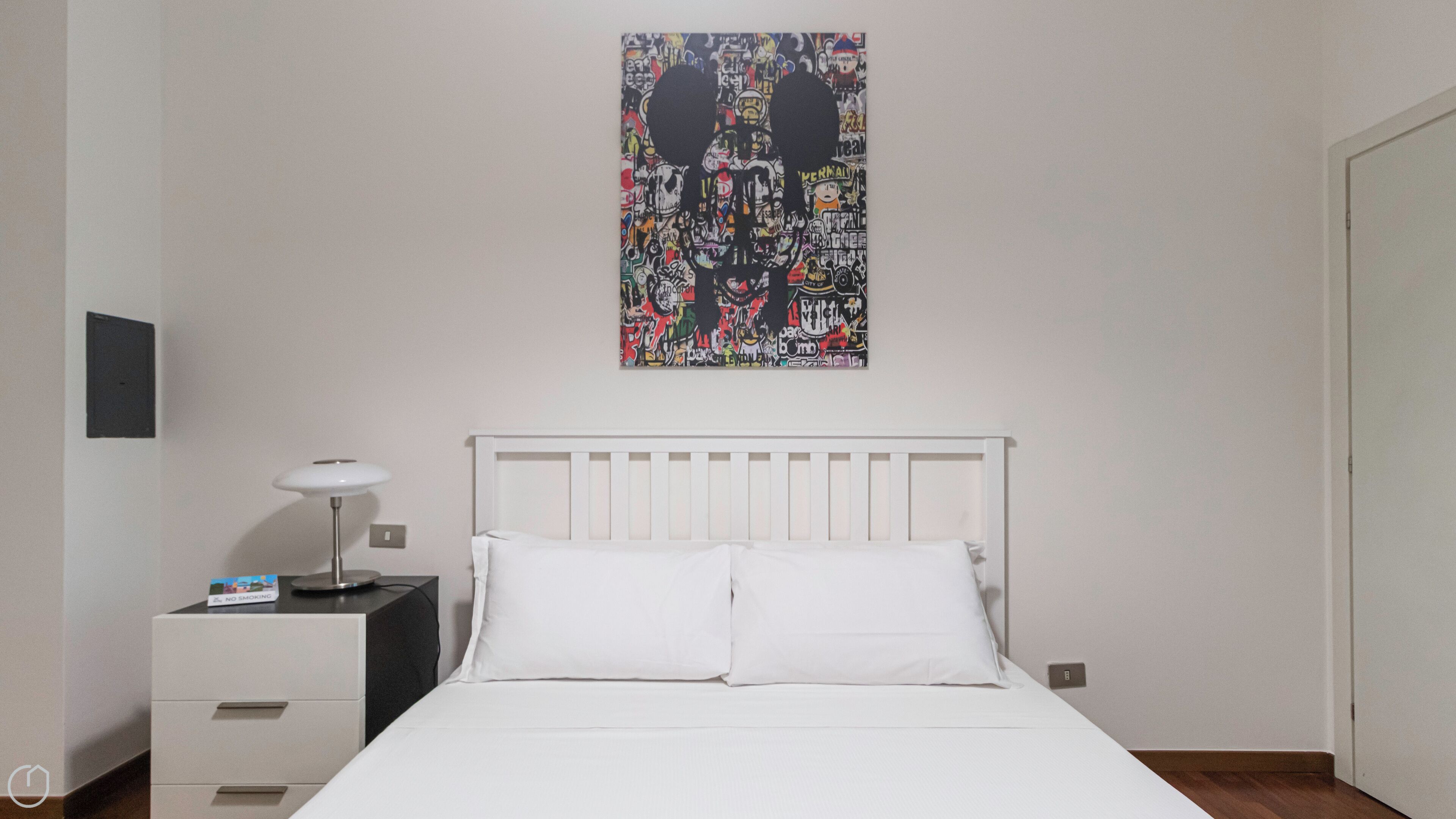 Een slaapkamer met een wit bed, witte kussens, een witte nachtkastje met een lamp, en een muur met een kleurrijk abstract schilderij.