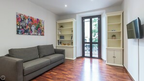A living room with a grey sofa, wooden flooring, a colorful abstract painting, built-in shelving with books and decorative items, and a TV mounted on the wall. - Italianway Easy - Buonarroti 5 (Milan)