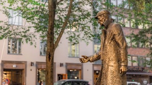 Una estatua de bronce de un hombre con traje y brazos extendidos, situada en un entorno urbano con edificios y árboles de fondo.