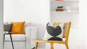 Un salón moderno con un sofá blanco, una silla de madera con cojines, una mesita con un libro y una estantería blanca con libros y un busto.