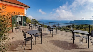 Een terras buiten met tafels en stoelen, met uitzicht op een prachtig zeegezicht en verre bergen.