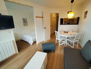 Een knus woongedeelte met een bed, een eettafel met stoelen, een bank, een televisie en een keuken met een hangende lamp.