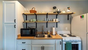 Cocina compacta con armarios blancos, un microondas, una estufa y repisas superiores con cristalería y una cesta.