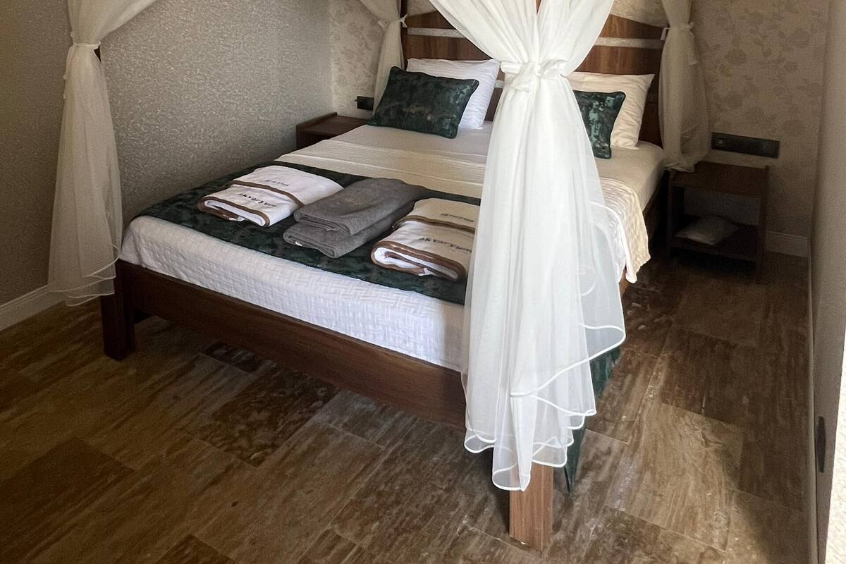 A four-poster bed with white drapes, two pillows, and neatly folded towels on a wooden floor.
