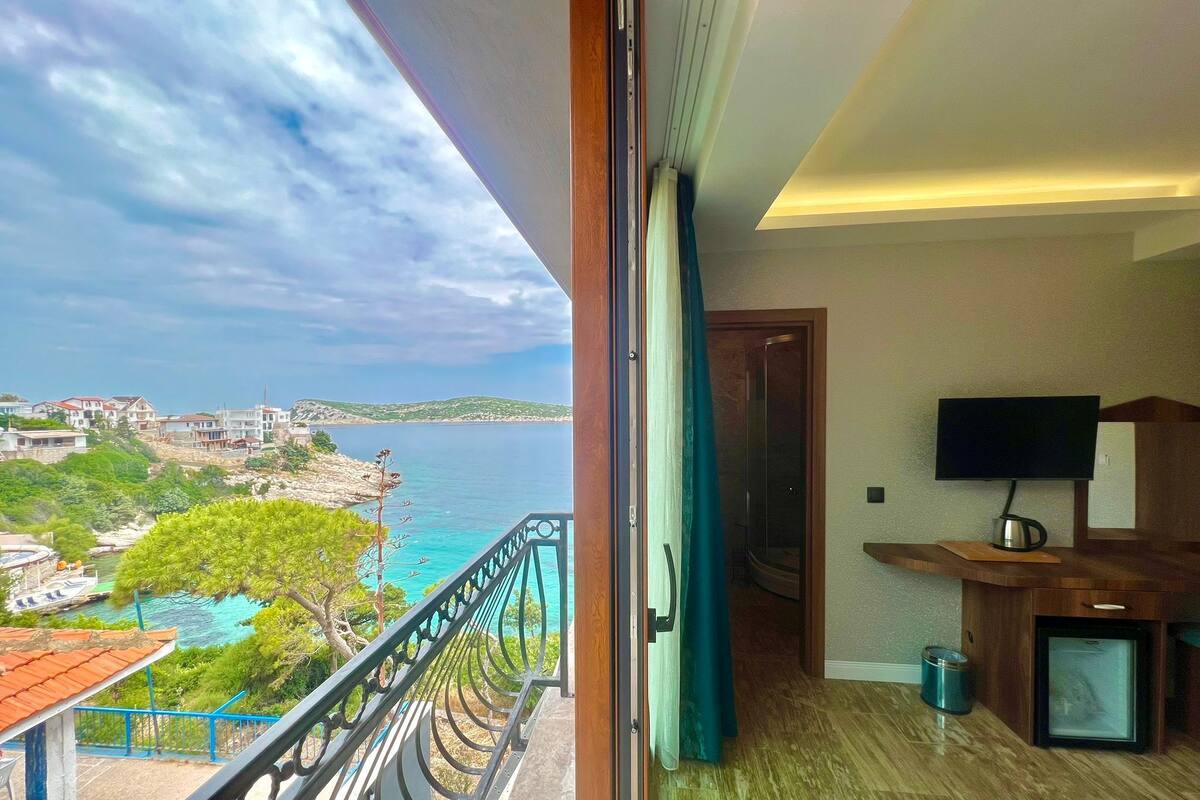 A balcony with a view of the sea and buildings, a TV, and a small desk with a cup.