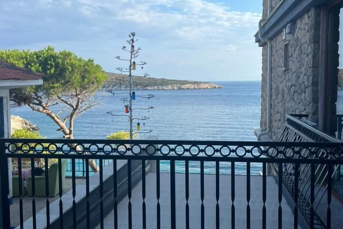 A balcony with a view of the sea, a stone building, and a tree.