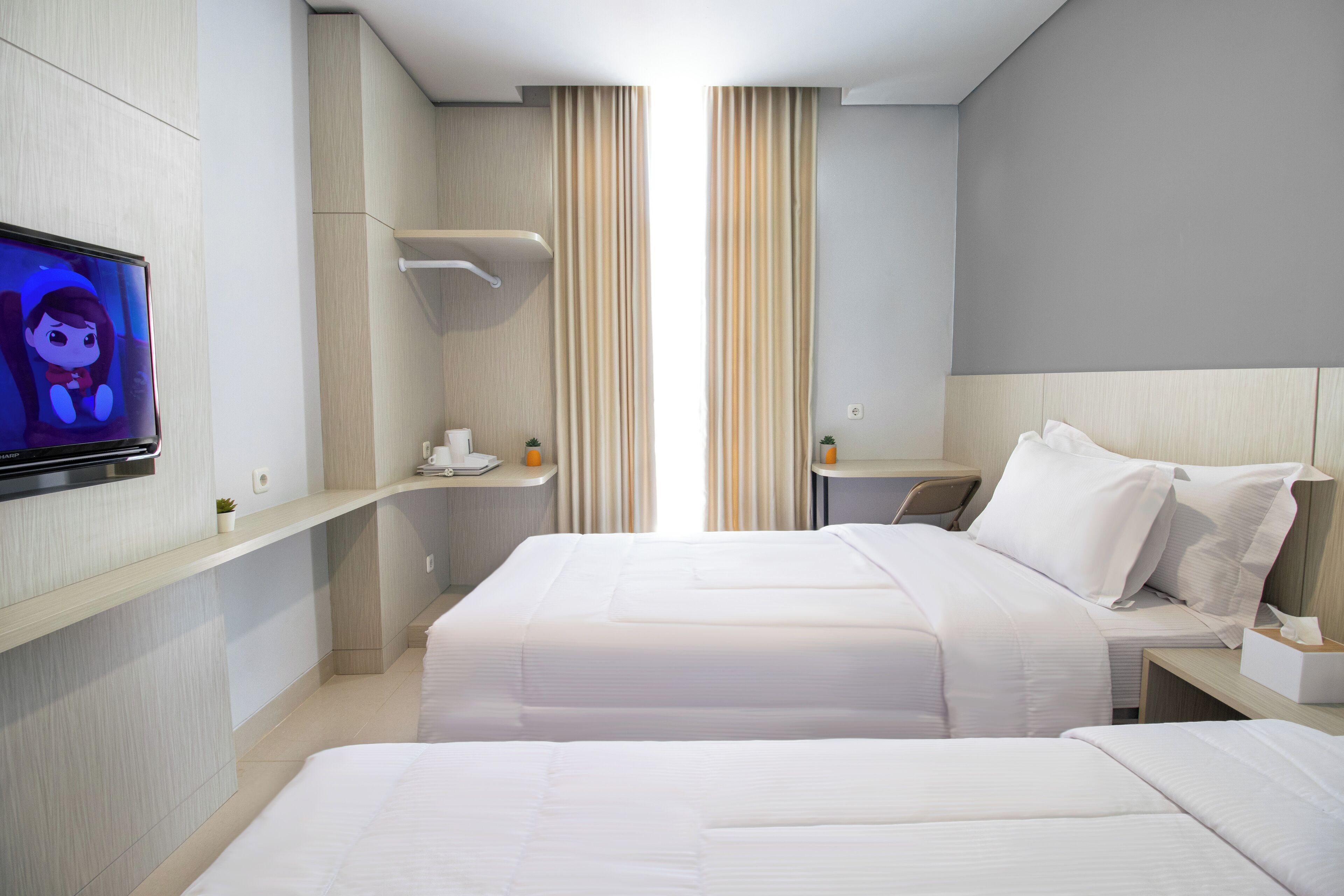 A modern hotel room with two beds, a TV on the wall, a small table, and a window with curtains.