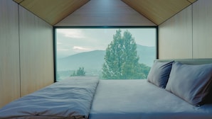 A bedroom with a large window, a bed, and a view of a tree and mountains. - Bobocabin Gunung Mas (Cisarua)