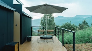 A wooden deck with a table and chairs, an umbrella, and a view of mountains. - Bobocabin Gunung Mas (Cisarua)