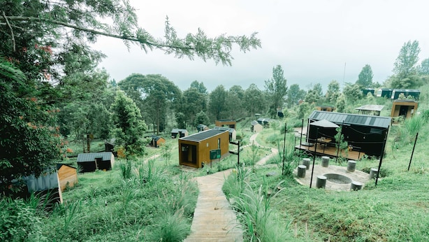 A wooden pathway leading through a lush, green landscape with multiple small buildings, some with solar panels, surrounded by trees and vegetation. - Bobocabin Gunung Mas (Cisarua)