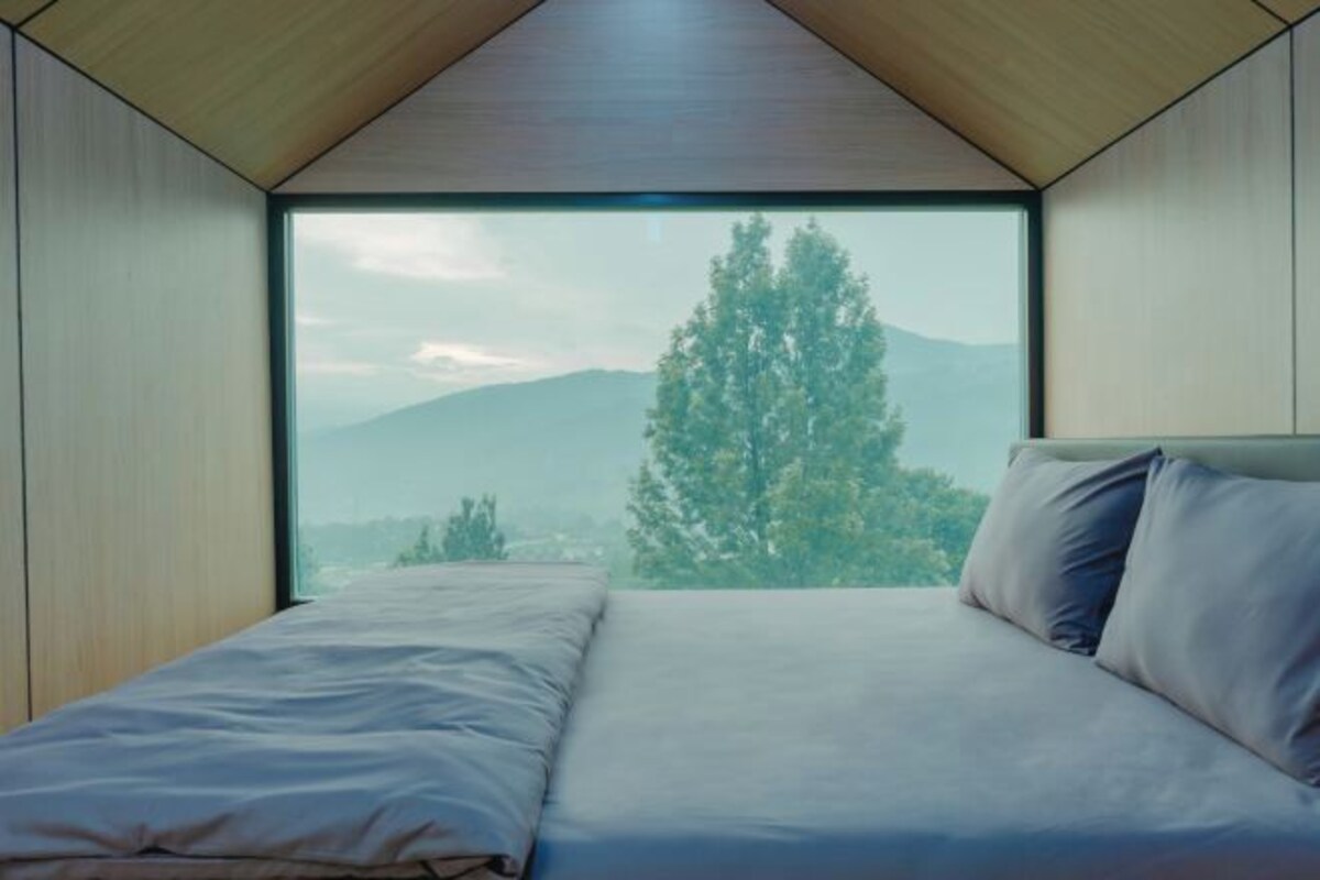 Un dormitorio con un ventanal grande, una cama y vistas a árboles y montañas.