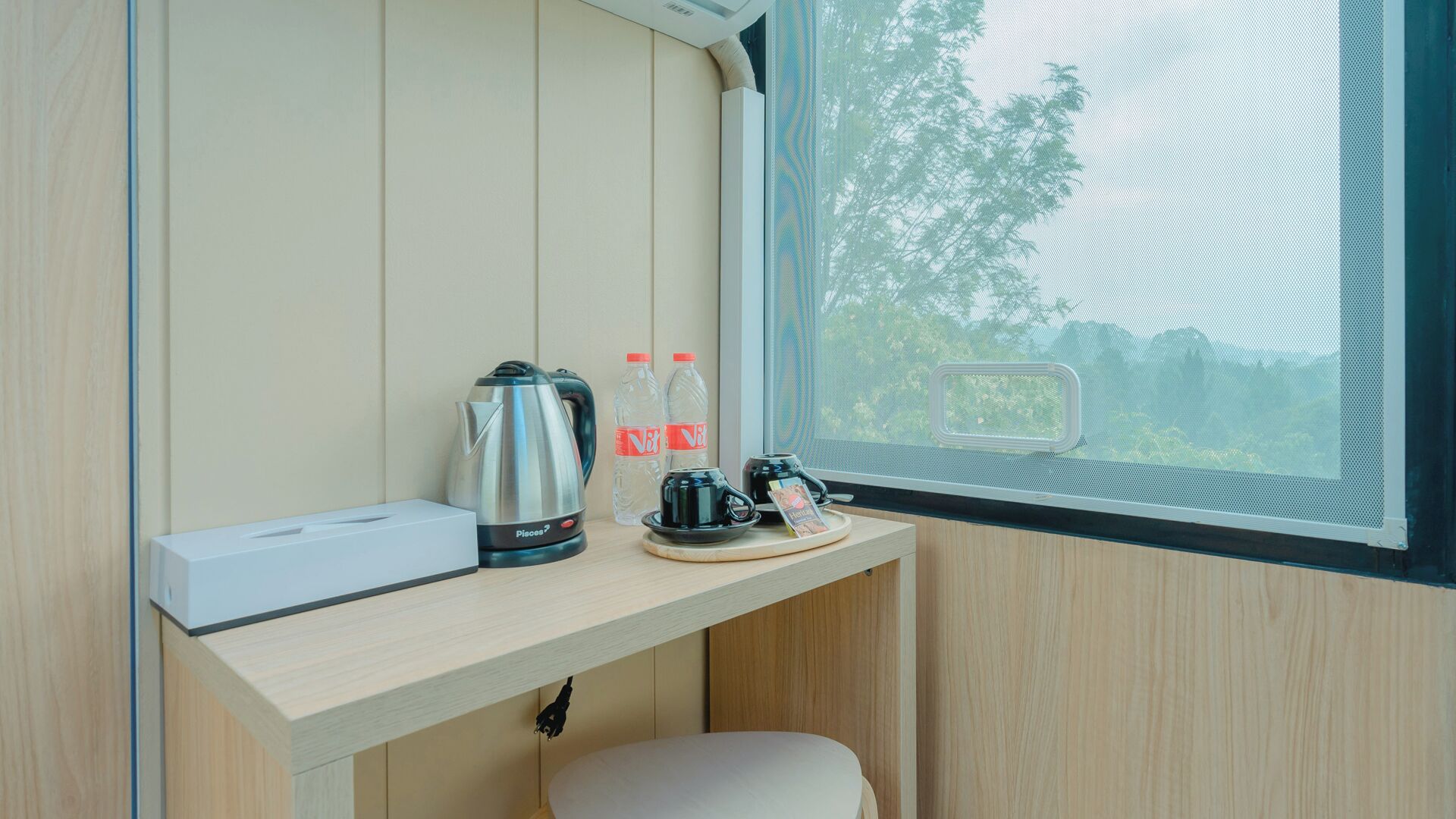 Un petit bureau en bois, sur lequel se trouvent une bouilloire, deux bouteilles d’eau et une tasse, est placé près d’une fenêtre donnant sur des arbres.