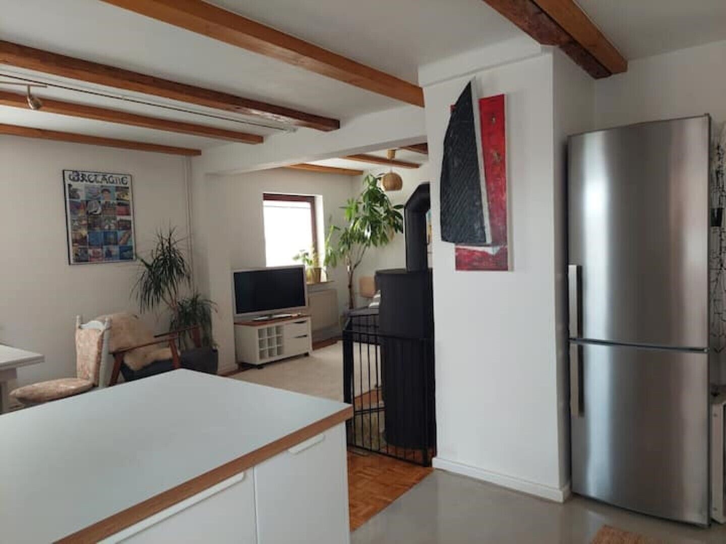 Eine moderne Küche mit einem großen, edlen Kühlschrank aus Edelstahl, Holzbalken an der Decke, einem Essbereich mit weißem Tisch und einem Wohnzimmer mit Fernseher und Pflanzen.