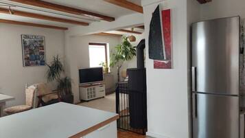 Eine moderne Küche mit einem großen, edlen Kühlschrank aus Edelstahl, Holzbalken an der Decke, einem Essbereich mit weißem Tisch und einem Wohnzimmer mit Fernseher und Pflanzen.