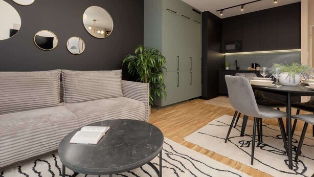 A modern living space with a sofa, a round coffee table, a dining table with chairs, and a kitchen area with cabinets and a microwave. - Stylish Apartment with Balcony | Air Conditioning and Parking (Warszawa)