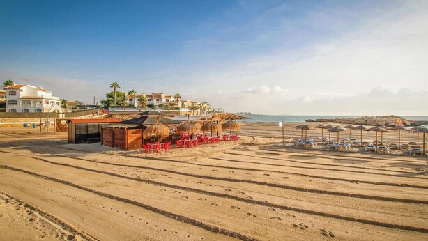 Una playa con filas de tumbonas y sombrillas, una pequeña cabaña con sillas rojas, y un edificio con balcones al fondo.