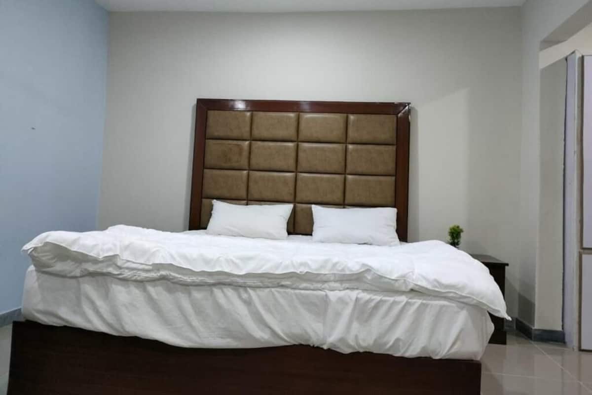 A neatly made bed with a wooden headboard and white bedding in a room with light-colored walls and a tiled floor.