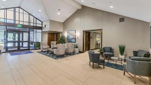 Een ruime lobby met een hoge plafond, grote ramen en diverse zithoeken, waaronder fauteuils en banken.
