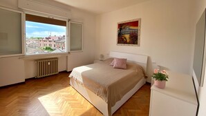 A bedroom with a large window, a radiator, a bed with a pink pillow, a painting on the wall, and a small plant on a nightstand. - BELLAVISTA, bright and stylish two bedroom apartment in the heart of Ravenna (Ravenna)