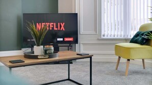 A living room with a television displaying 'NETFLIX,' a small table with a plant, a yellow chair, and a window with blinds. - Spacious 4BR retreat Eden Vale | by Stay With Us (Sunderland)