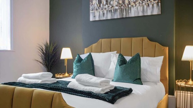 A neatly made bed with white and teal pillows, a folded white towel, and a dark green throw blanket. There are two bedside lamps on either side of the bed. - Spacious 4BR retreat Eden Vale | by Stay With Us (Sunderland)