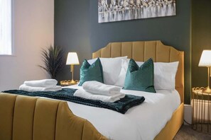 A neatly made bed with white and teal pillows, a folded white towel, and a dark green throw blanket. There are two bedside lamps on either side of the bed. - Spacious 4BR retreat Eden Vale | by Stay With Us (Sunderland)