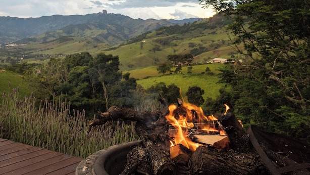 Un pozo de fuego de piedra con fuego encendido, rodeado por una terraza de madera y con vistas a un paisaje pintoresco con colinas y árboles.