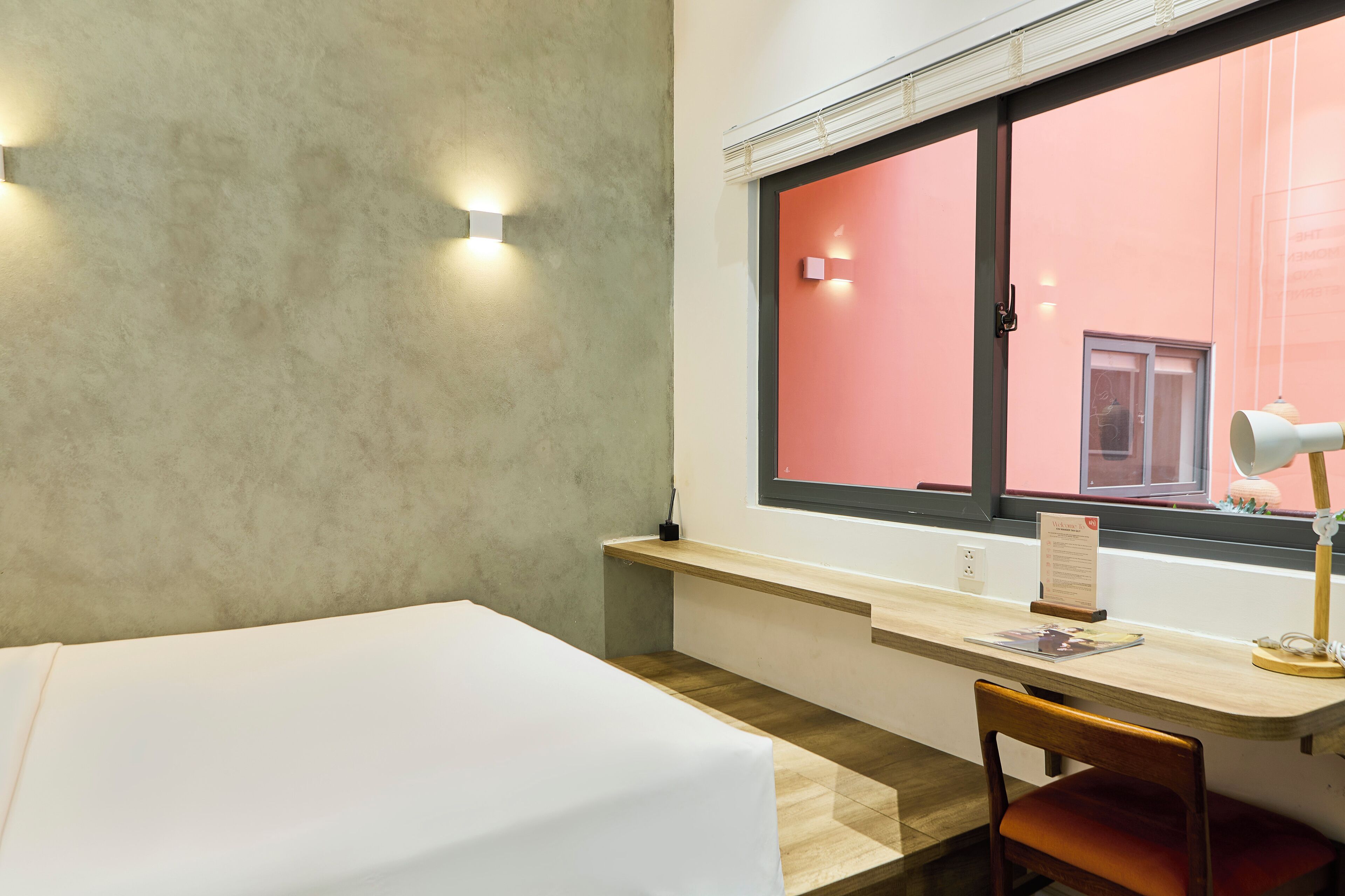 A hotel room with a bed, a desk, a chair, a window with blinds, and a desk lamp.