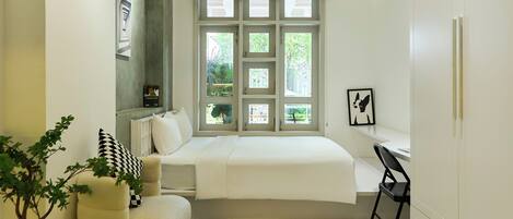 A bedroom with a bed, a desk, a chair, a window with multiple panes, and a plant.