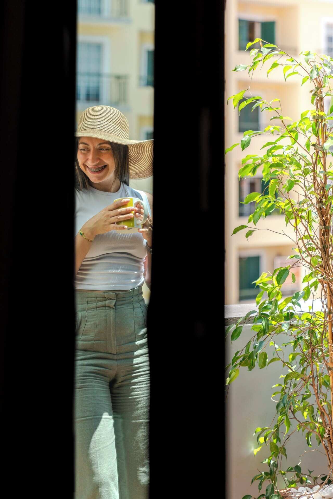 A person standing in a doorway, holding a drink, with a potted plant and a building in the background.