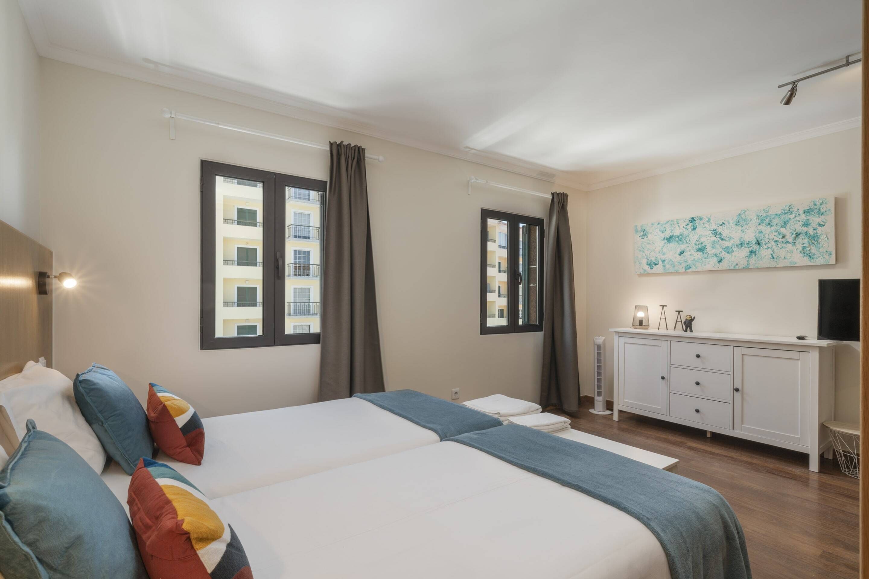 Ein Hotelzimmer mit zwei Betten, einer weißen Kommode, einem Bild an der Wand und einem Fenster mit Vorhängen.