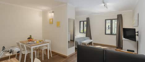 Una sala de estar moderna con un sofá negro, una mesa de comedor con sillas, un televisor montado en la pared y un ventanal con cortinas.