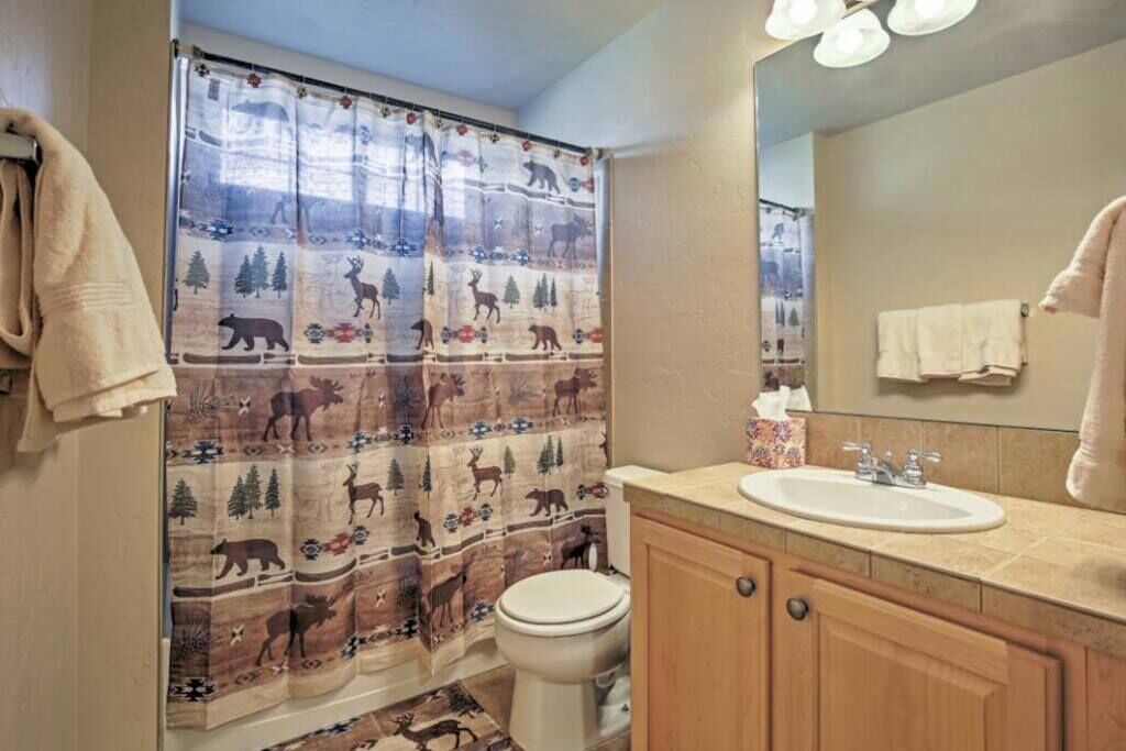A bathroom with a shower curtain featuring a forest and wildlife print, a white toilet, a sink with a mirror above it, and wooden cabinets.