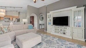 A living room with a sectional sofa, ottoman, TV, and white cabinetry. - Serenity Haven with Private Pool in Gilbert (Gilbert)