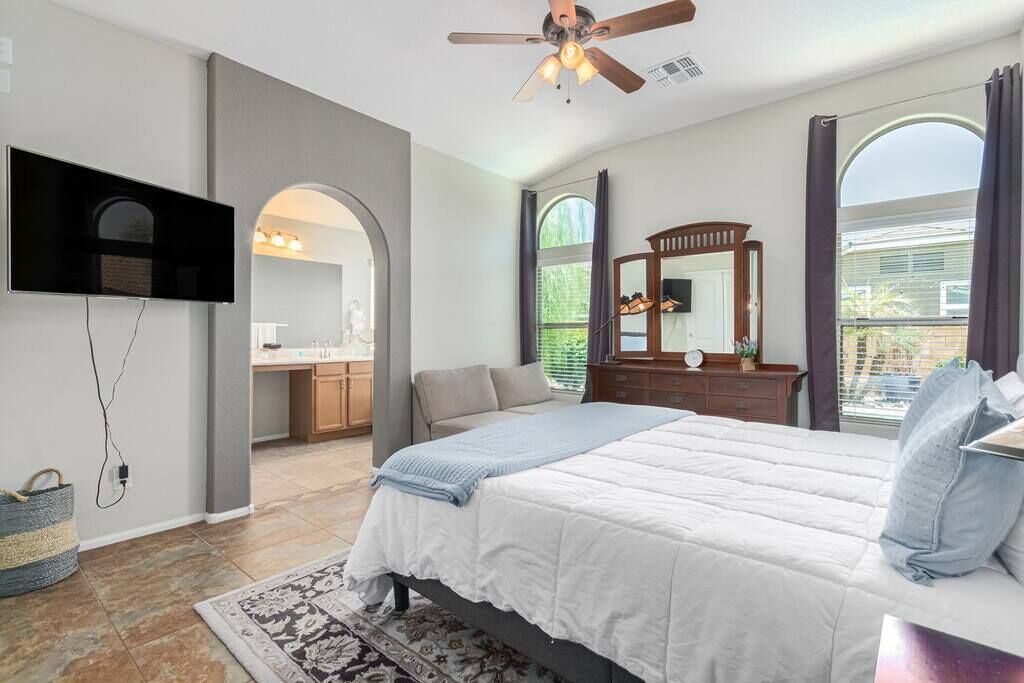 A bedroom with a bed, a ceiling fan, a TV mounted on the wall, a dresser, and a window with curtains.