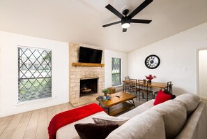 A living room with a stone fireplace, a ceiling fan, a dining table with chairs, a sofa, and a wall clock.