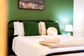 A neatly made bed with white and green pillows, a rolled towel, and a bedside lamp.