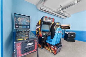 Une salle avec des bornes d’arcade, dont un jeu Spider-Man et un jeu de course, avec un mur bleu et une grille d’aération au plafond.