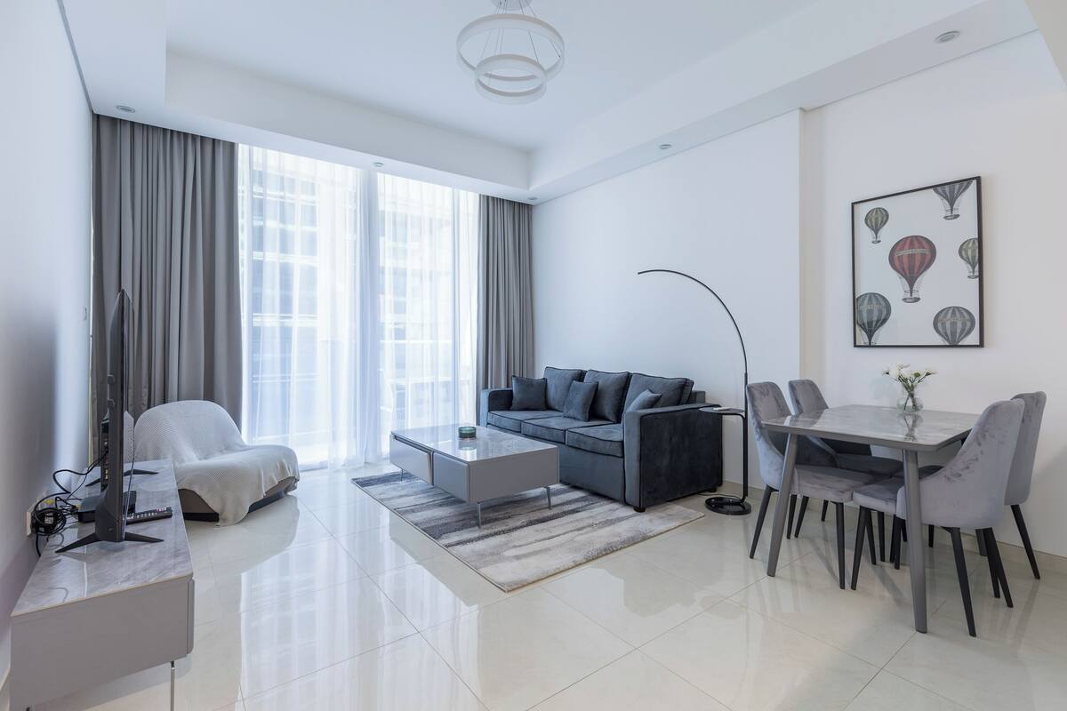 A modern living room with a grey sofa, a coffee table, a dining table with chairs, and a large window with curtains.