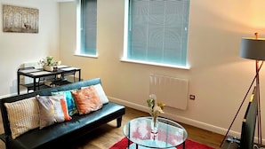 A modern living room with a black leather sofa, a glass coffee table, and a red rug. There are two windows with blinds, a radiator, and a painting on the wall. - Captivating 2-bed Apartment in Bedford (Bedford)