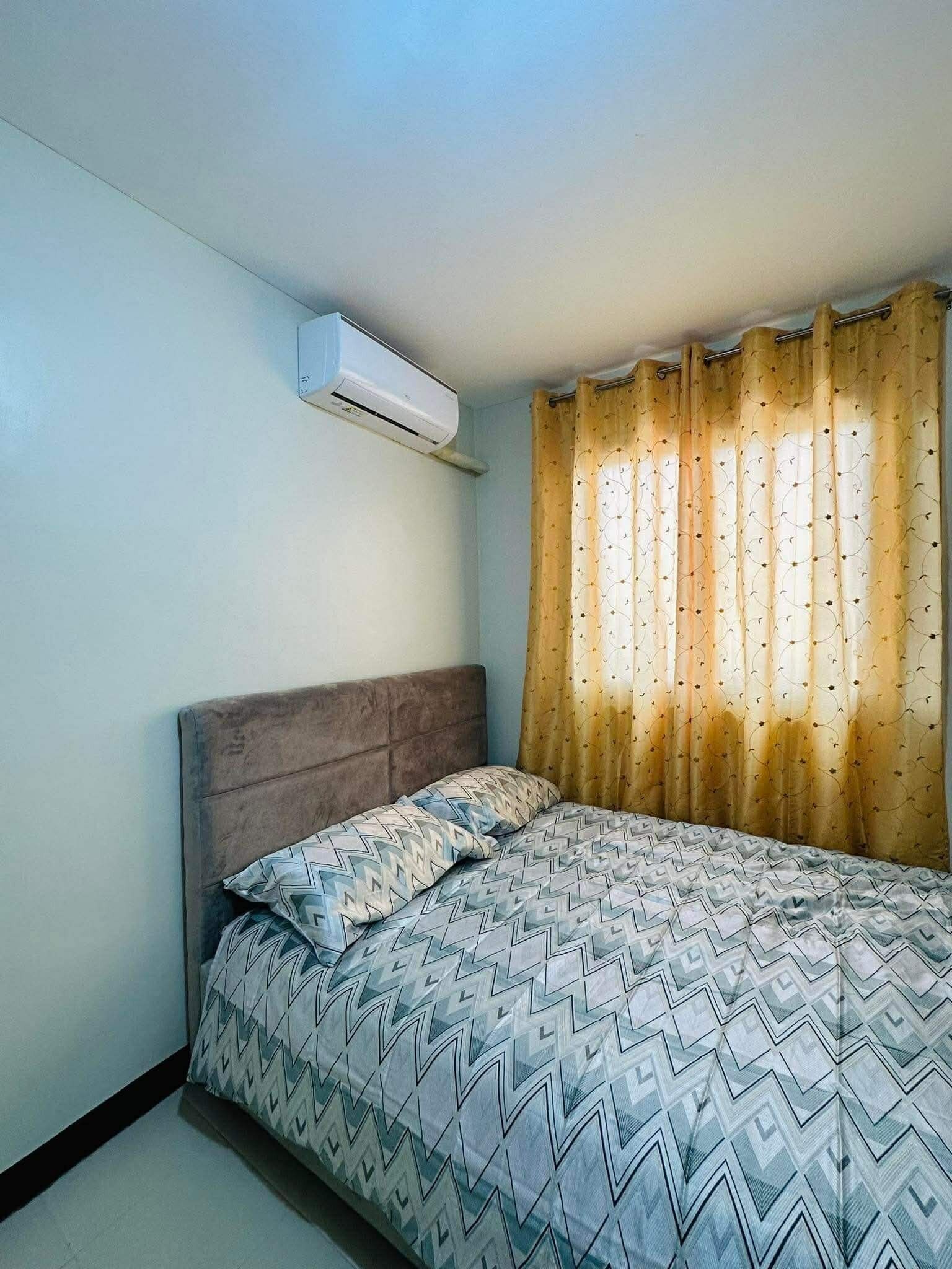 A bedroom with a bed, a headboard, an air conditioning unit, and a window with patterned curtains.