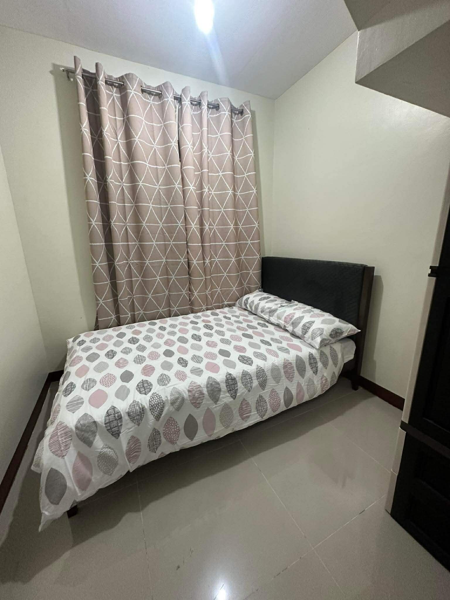 A single bed with a patterned bedspread, a headboard, and a curtain with a geometric design.