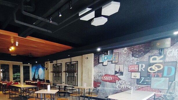 Un café moderno con varias mesas y sillas, un mural con distintas frases y un techo de madera.