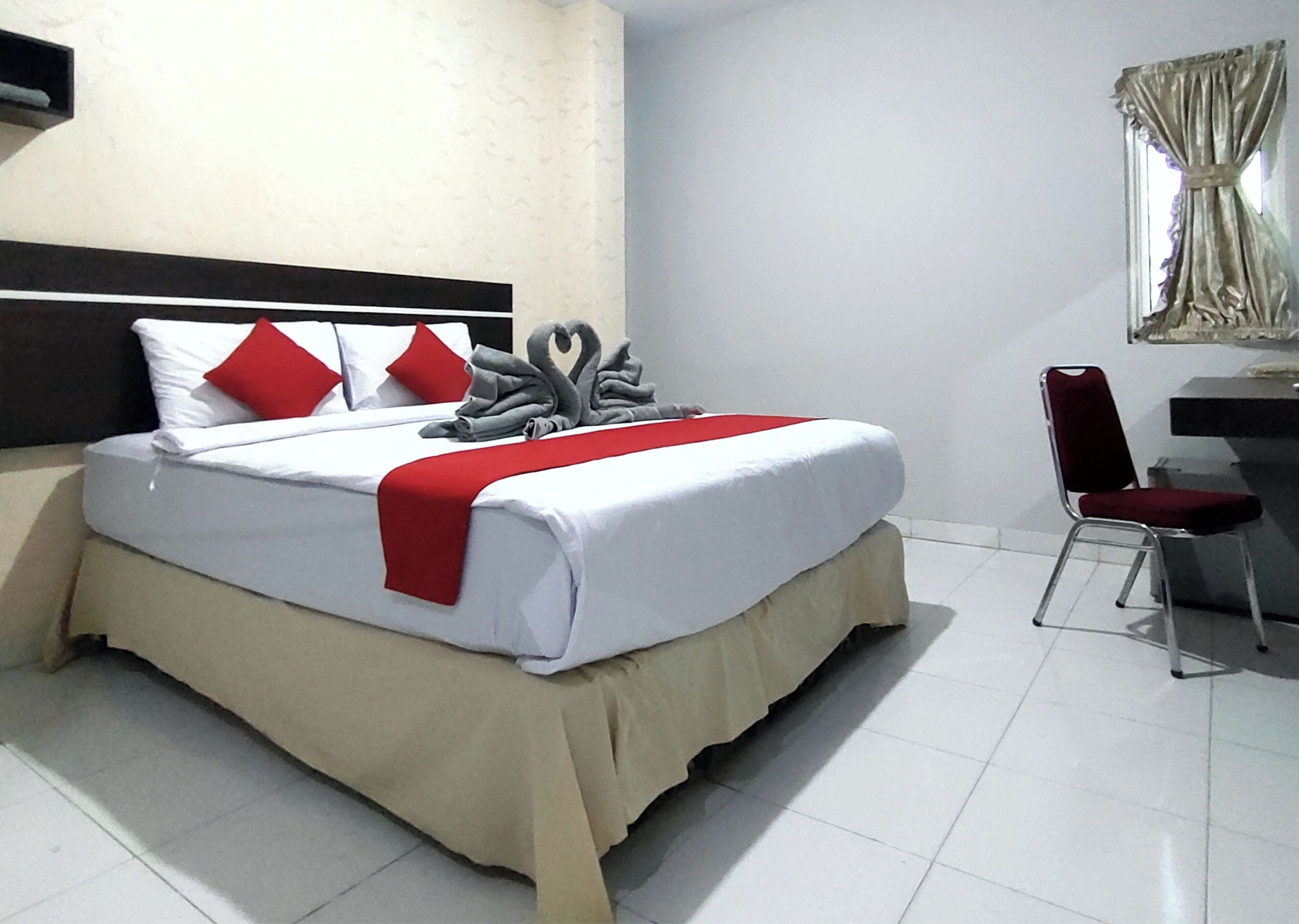 A neatly made bed with white bedding, red pillows, and a red blanket. There is a chair and a desk with a mirror in the room.