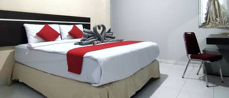 Una cama bien hecha con ropa blanca, almohadas rojas y una manta roja. Hay una silla y un escritorio con un espejo en la habitación.