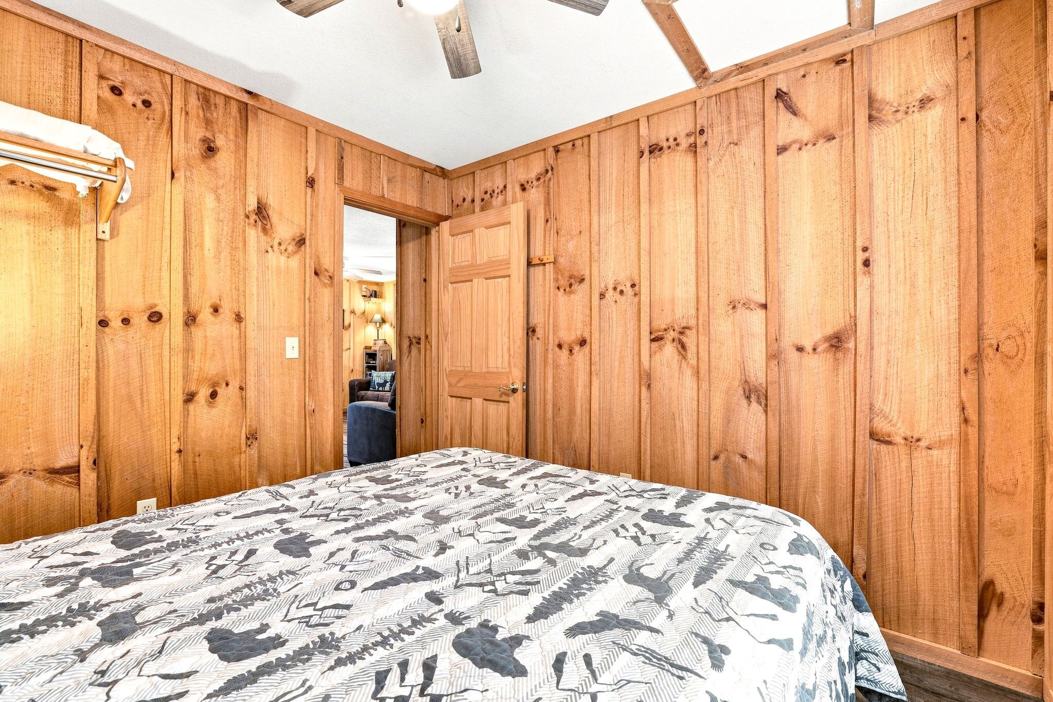 A room with wooden panel walls, a bed with a patterned bedspread, a ceiling fan, and a visible doorway leading to another room.