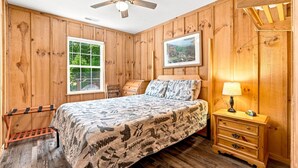 A bedroom with a bed, a nightstand, a lamp, a ceiling fan, and wooden walls. - Cabin the the Gap- Motorcycle Friendly with Pond (Bryson City)
