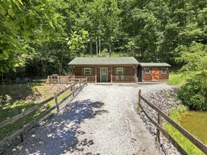 A cabin with a gravel driveway leading to a wooden bridge, surrounded by dense forest.