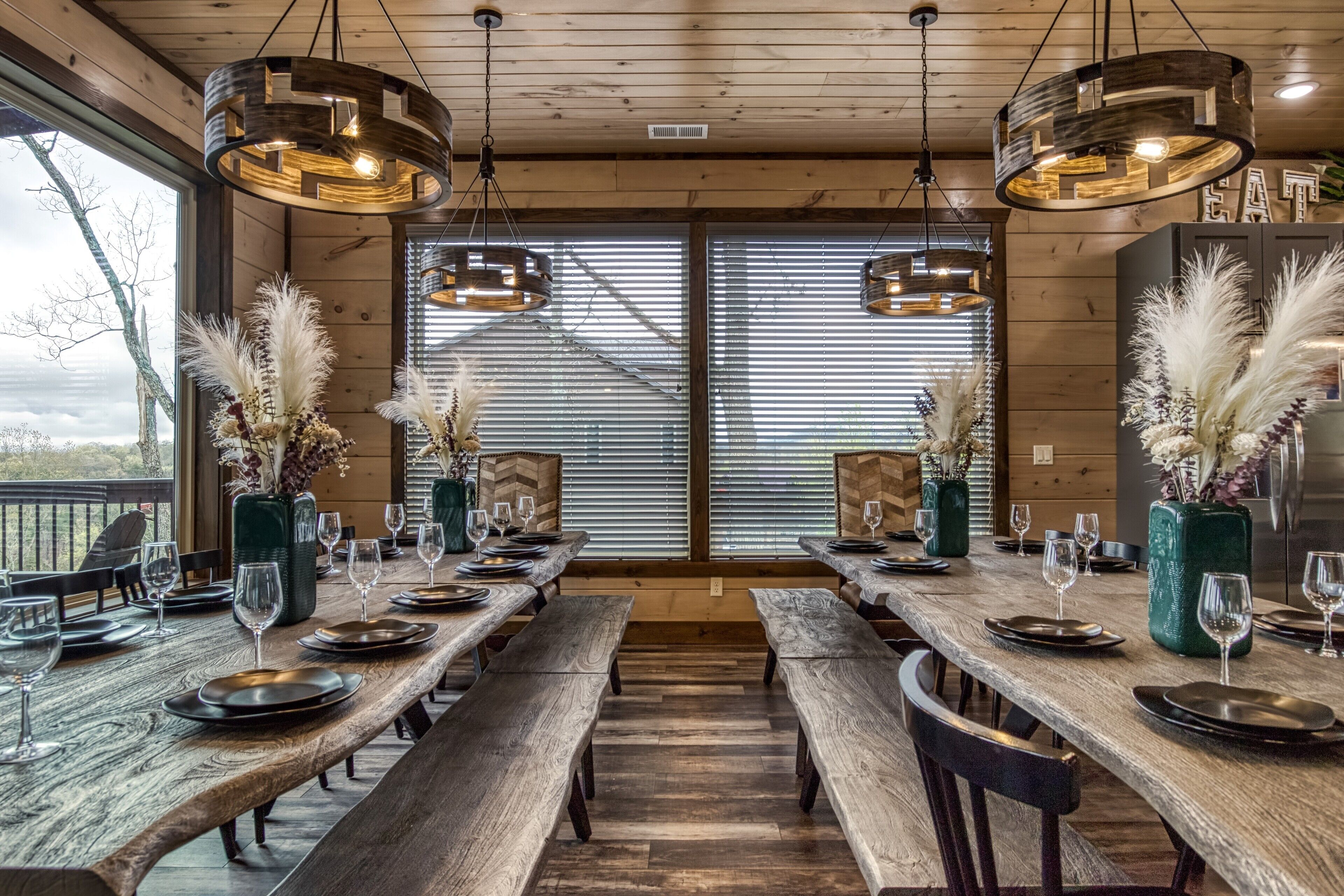 A long wooden dining table set with plates, glasses, and benches, with hanging pendant lights and decorative vases.