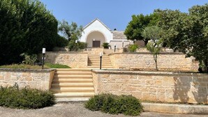 Un bâtiment en pierre doté d’une entrée centrale en arc de cercle, entouré de verdure et accessible par un escalier en pierre.